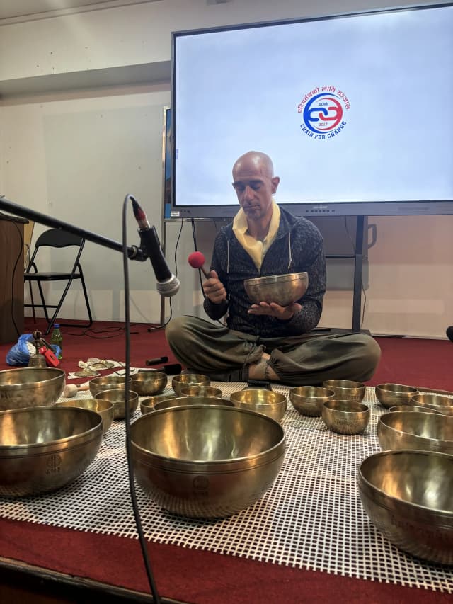 Singing Bowl Master Mr. Oscar is holding a bowl and playing. In front of him, there are many singing bowls placed and in the background logo of CFC is displayed on the screen.