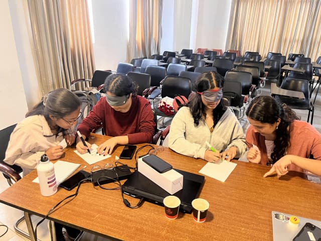 Two of the participants are being blindfolded and are drawing on a paper as per the instruction given by their partners.