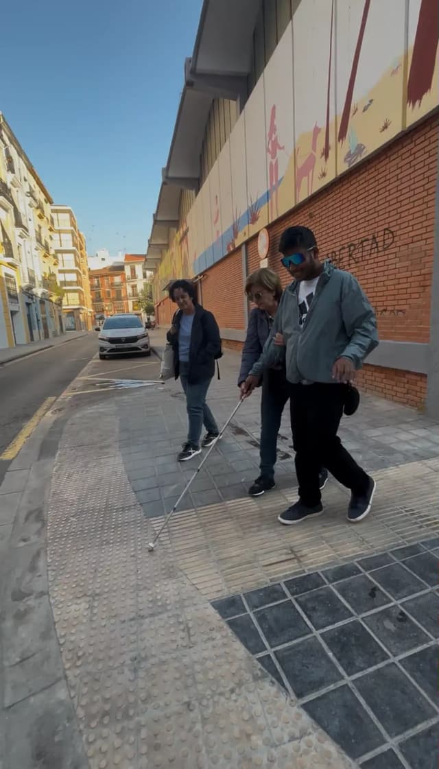 Mati Barbara who worked as the Técnico Rehabilitación at ONCE is instructing our Founder Abhishek Shahi to use the white cane in the road of Valencia, Spain. Fanny Collado is observing them. In the background, there is a car and some buildings.