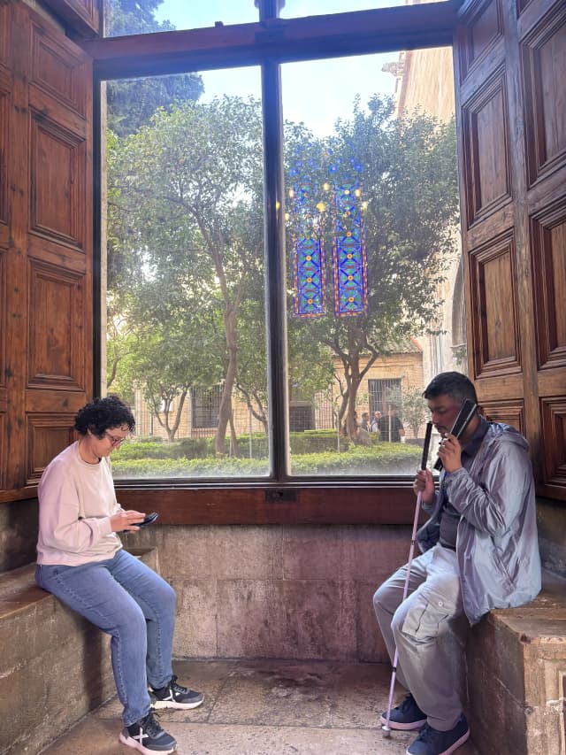 A picture taken in front of a big window of La Lonja, where our Founder Abhishek Shahi is listening to the audio device and Fanny Collado is using her phone.